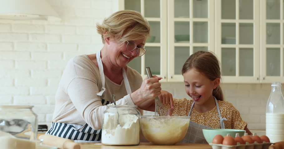 60s granny teach little granddaughter to cook, mixing dough mixture in bowl enjoy process and communicating together in cozy kitchen. Home hobby, older gen share skill with younger generation concept | Shutterstock HD Video #1070888152