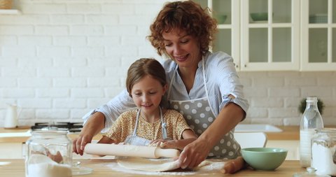 Mom and little daughter wear aprons rolls out the dough for baking cooking together in kitchen. Woman teach kid use rolling-pin flattening dough preparing home-made pastry for family. Cookery concept