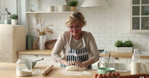 Elderly woman 60s older granny wear apron and glasses kneading dough with hands for pastries making pizza or pie cooking in modern kitchen. Family recipe, routine, home hobby, cookery process concept