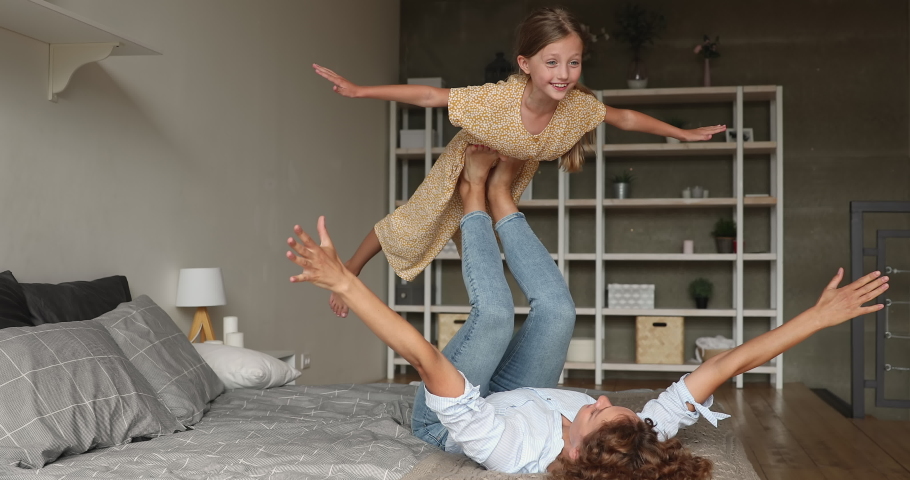 In cozy bedroom active strong mother lying on comfy bed holding lifting small daughter on legs, child spread arms like plane wings imagines like fly in air. Sportive family, yoga practice, fun concept Royalty-Free Stock Footage #1070890873