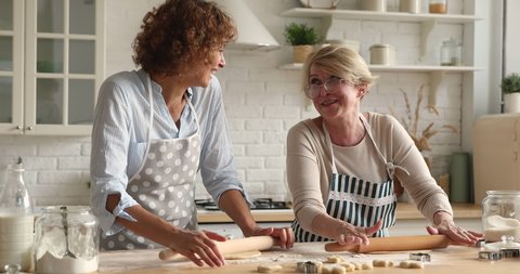 Cheerful older woman her grown up adult daughter talk make flattening dough with rolling-pins in kitchen. Family use ecological products high quality ingredients cooking together laughing feel happy