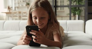 Close up pretty little adorable girl resting at home with smartphone, lying indoor holding in hands gadget watch videos, play games online enjoy modern wireless technology and internet usage concept