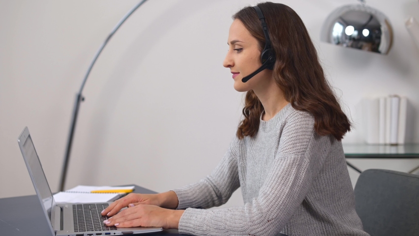 Smiling young saleswoman in a headset is holding the phone conversation, female employee involved video conference, customer service representative sitting in front of a laptop and talks, side view Royalty-Free Stock Footage #1070897809