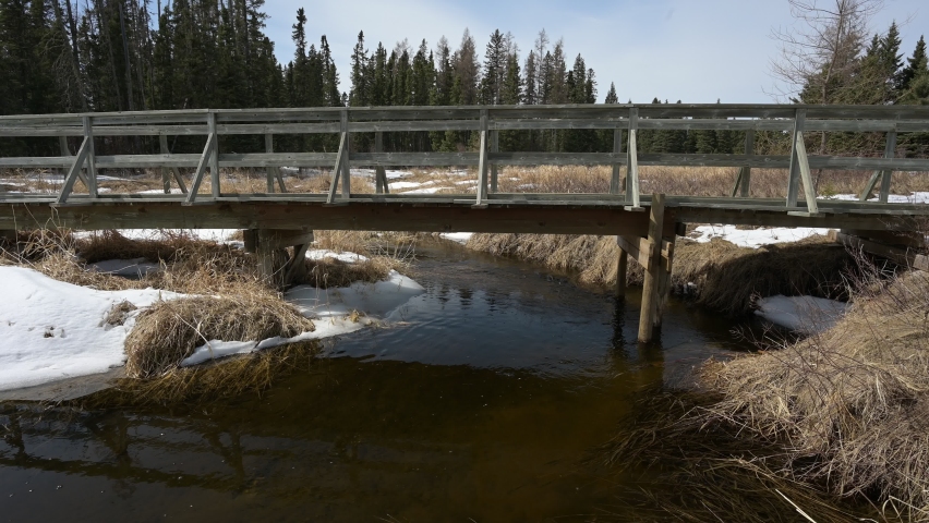 A small stream with clear slightly brown stained water runs under an old walking bridge and through a meadow of dead grass with a boreal 
