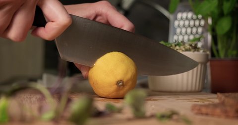 Close up side view of woman slicing fresh ripe lemon on wooden cutting board with big kitchen knife.