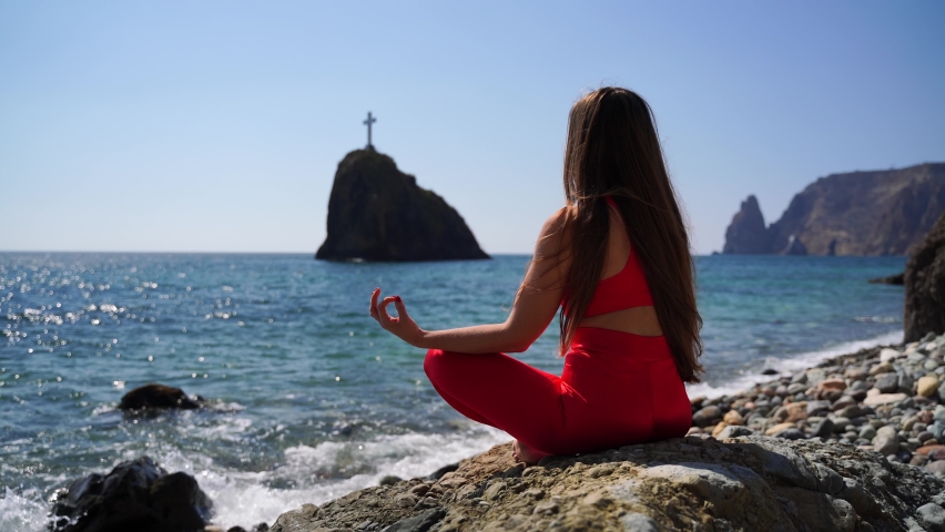 A young woman in red sportswear relaxes while practicing yoga on the beach by the calm sea, close-up of hands, gyan mudra and lotus pose.
