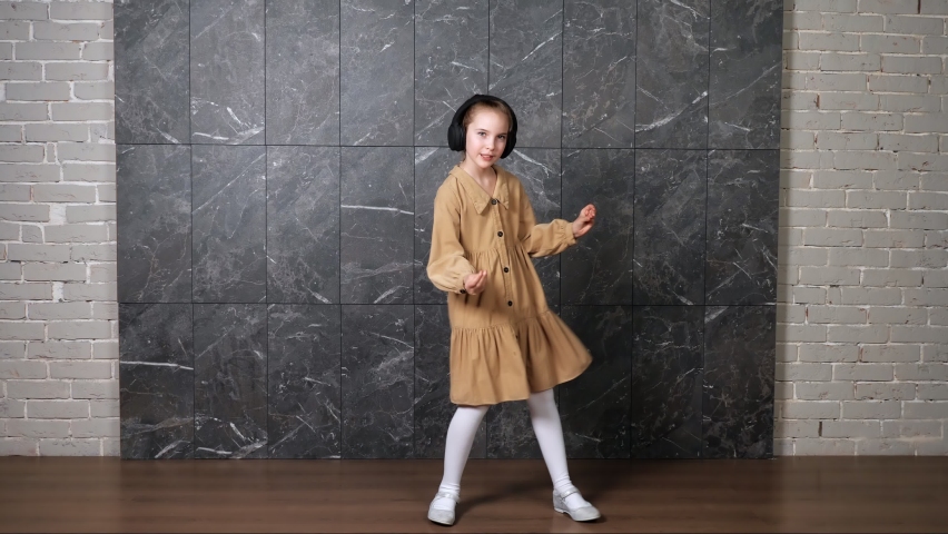 Delighted active girl in brown dress dances wearing black headphones against designed marble wall surrounded by grey bricks in hallway