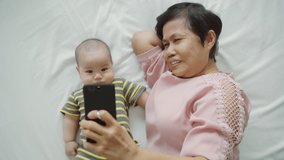 Happy Asian Grandmother and little baby boy lying on bed taking selfie photos with mobile phone. Senior woman and Grandchild or Grandson doing video call on smartphone with family together