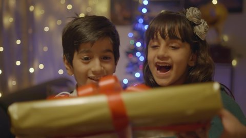 Two well dressed adorable young kids are smiling and receiving their Christmas present saying thank you in a decorated house. Happy and elated cute girl and boy accepting the gift box from parents 
