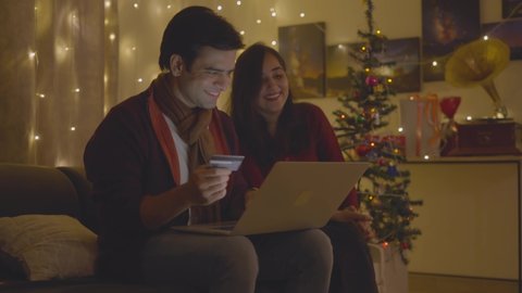 A shot of a young attractive Indian Asian well-dressed married couple sitting at home on a couch doing online shopping on a laptop using credit or debit card during the occasion of Christmas eve 