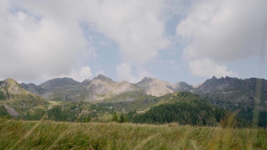 Static shot of male hiker with trekking poles walking along rural path during sunny day with clouds and beautiful mountain range in background. Trentino,Italy.