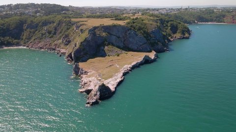 Aerial pan shot of famous Long Quarry Point on high hill surrounded by green blue ocean water in sun. Torquay,England.