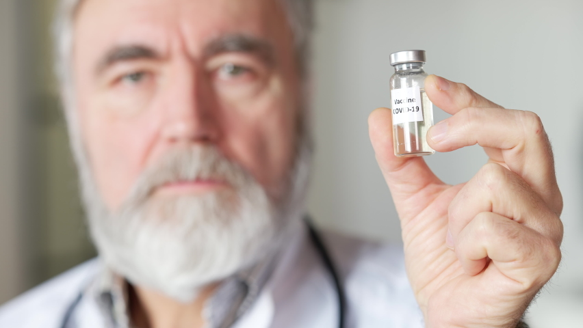 Close-up of Covid-19 vaccine jar in male hand with blurred senior doctor at background. Confident serious Caucasian man posing with coronavirus medication indoors. Pandemic treatment Royalty-Free Stock Footage #1070940742