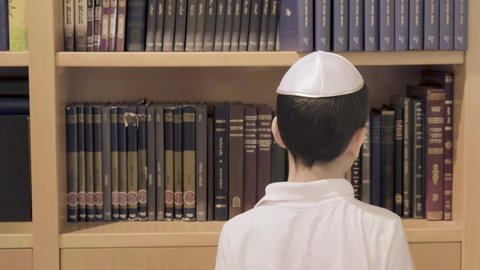 Jewish boy wearing Kippah Yarmulke and Talit, looking at religion Torah bible books. Mitzvah celebrating ceremony in the synagogue. Holy Jewish books on shelves.  Ramat Hasharon, Israel – April 17, 20