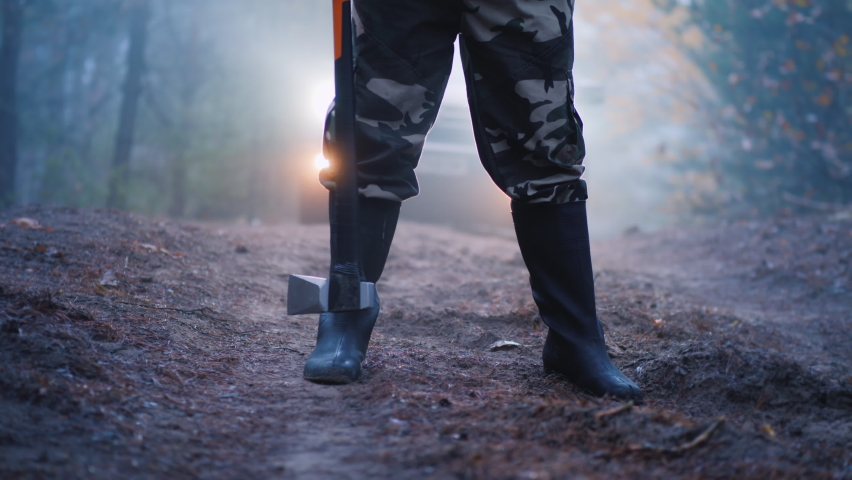 Legs in boots of a man who holds an ax, car headlights shine from behind