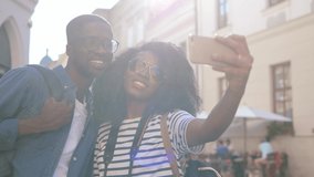 Young happy african american couple of tourists standing on street in sunlight and recording short video for social networks on smartphone.