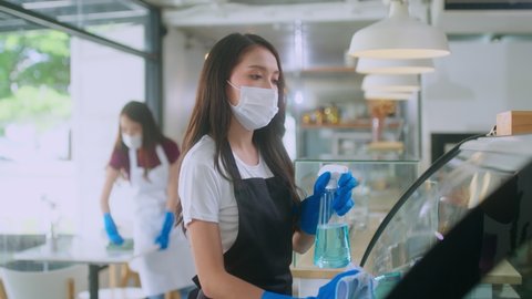 Young Asian female worker woman in mask rubber glova and apron uniform cleaning and preparing cafe counter for customer in coffee shop, sanitizer disinfection during coronavirus Covid-19 pandemic