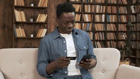 Cheerful african man in casual clothing with smartphone make online payment shopping with credit card sitting on sofa at home paying for purchases store buy