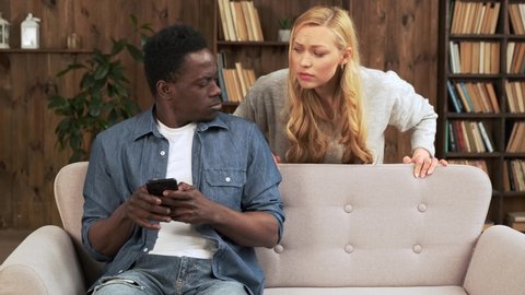 Cheater man dating online with a smart phone and girlfriend is spying sitting on a sofa at home. Mixed race ethnicity couple