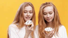 Sweet tooth family. Two cheerful redhead twin sisters eating sweet glazed donuts, enjoying pastry over orange studio background, slow motion