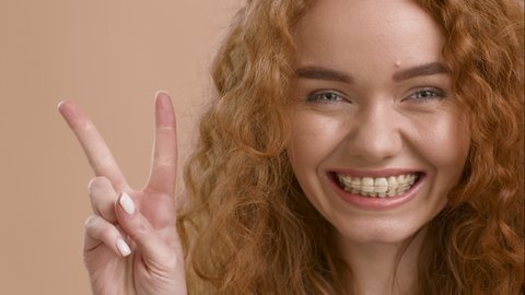 Red-Haired Millennial Woman Gesturing Victory And Peace Sign Smiling To Camera Posing On Beige Background. Studio Portrait Shot Of Cheerful Young Lady Showing V-Sign
