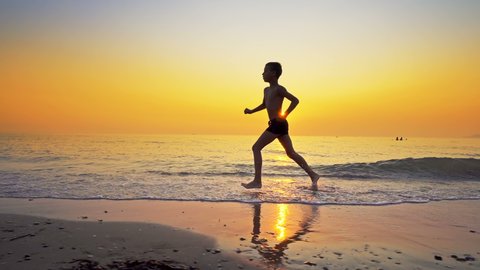 Closeup of young boy running, jogging on sea beach against sunset. Steadicam shot