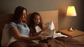 Young mother and adorable daughter flip through pages of children book together, choosing bedtime story while lying in bed in dark nursery bedroom before bedtime, near lamp. Shooting in slow motion. - Powered by Shutterstock - Get 15% off with code: PIKWIZARD15