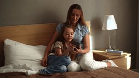 Portrait of happy young mother hugging to cute daughter together watching fun video on mobile phone, chatting on video call sitting on bed in bedroom of home. Shooting in slow motion.