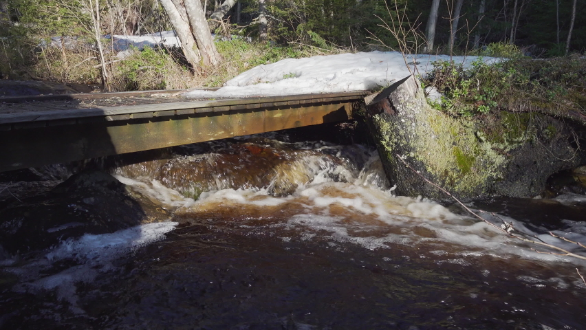 Very small wooden flat bridge over forest creek with much water flowing under it. Melting snow in forest close to the running water. Sweden, Europe. Original sound of running water