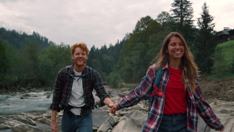 Positive couple hiking with backpacks along mountain river. Smiling travellers walking on rocky shore of river. Happy guy and girl holding hands. Young hikers exploring nature 