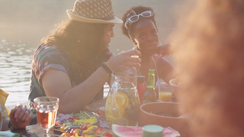 Young middle eastern man and Afro-American woman sitting at dinner table with friends, smiling and chatting during lake party on summer day