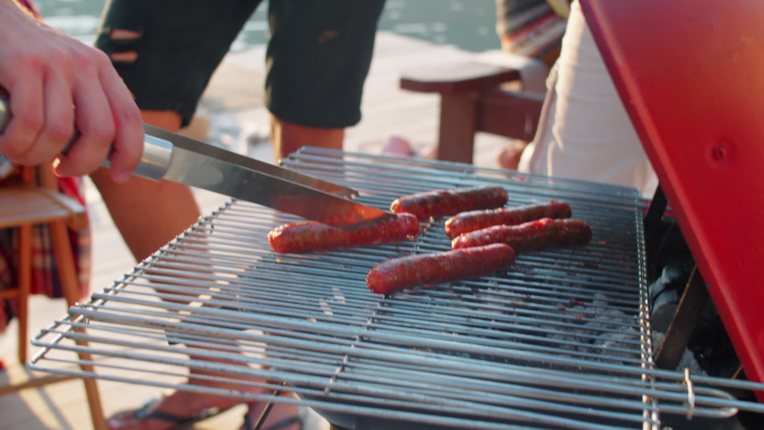 Tilt up shot of man taking bbq sausage from grill with metal tongues and putting it into bun while making hot dogs with help of friends on summer lake party Royalty-Free Stock Footage #1070978461
