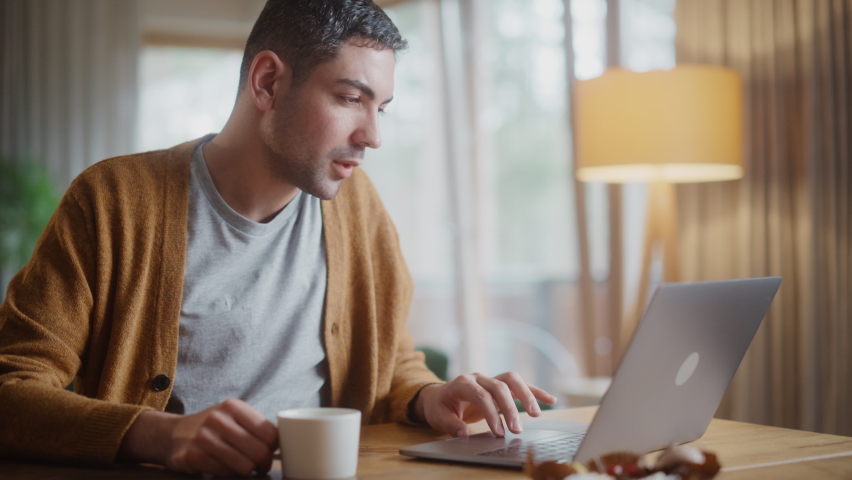 Handsome Adult Man Using Laptop Computer, Sitting in Living Room and Drinking Tea or Coffee in Apartment. Attractive Man is Online Shopping on Internet, Watching Funny Videos on Streaming Service. Royalty-Free Stock Footage #1070981476