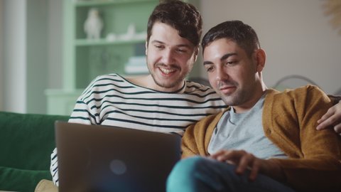 Portrait of Gentle Gay Couple Using Laptop Computer, while Sitting on a Couch in Cozy Stylish Apartment. Adult Boyfriends Online Shopping on Internet, Watching Funny Videos on Streaming Service.