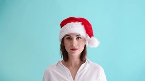 Beautiful caucasian woman wearing red santa hat and white shirt smiles and shows OK hand sign. Blue background. Studio shot. 4K resolution video. Christmas hand gesture theme.