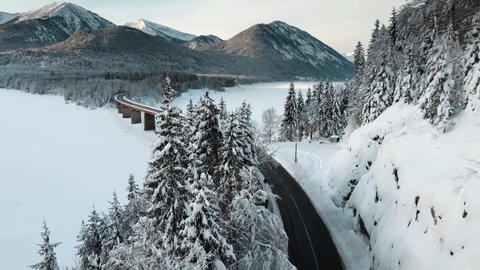 Aerial flying over snowy pine trees towards a majestic frozen lake and bridge on a mountain valley