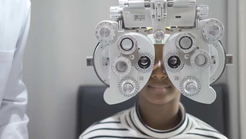 Optician doing optometry eye exam for black african american teen girl patient. Male optometrist with phoropter while examining patient. Royalty-Free Stock Footage #1070986996