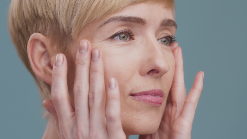 Anti aging skincare. Close up portrait of mature woman making facial massage, moisturizing her face skin, blue studio background | Shutterstock HD Video #1071009817
