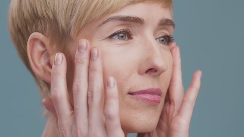 Anti aging skincare. Close up portrait of mature woman making facial massage, moisturizing her face skin, blue studio background