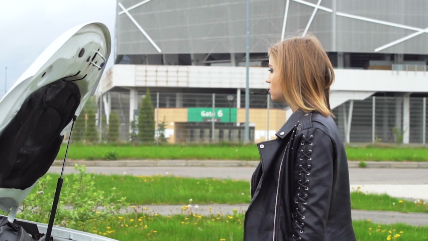 Stylish girl near her broken white car. Beautiful girl opens the hood of her new, white car. The girl diagnoses the breakdown of her car. A girl near the stadium looks under the hood of her broken car