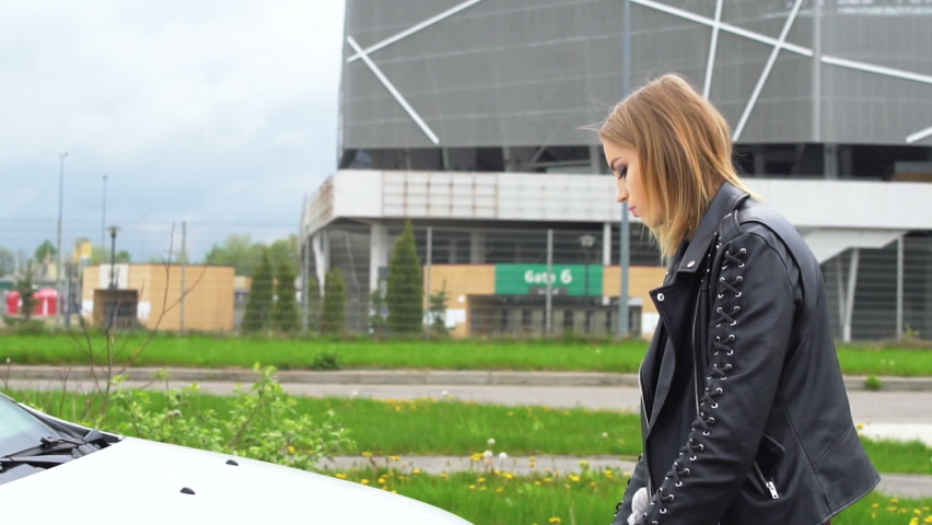 Stylish girl near her broken white car. Beautiful girl opens the hood of her new, white car. The girl diagnoses the breakdown of her car. A girl near the stadium looks under the hood of her broken car