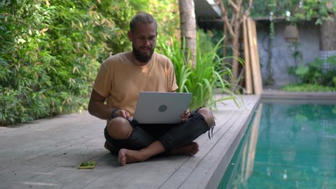 Caucasian digital nomad connecting to villa internet for doing remote work at pool terrace, skilled male freelancer reading web publication while making online booking via application on netbook