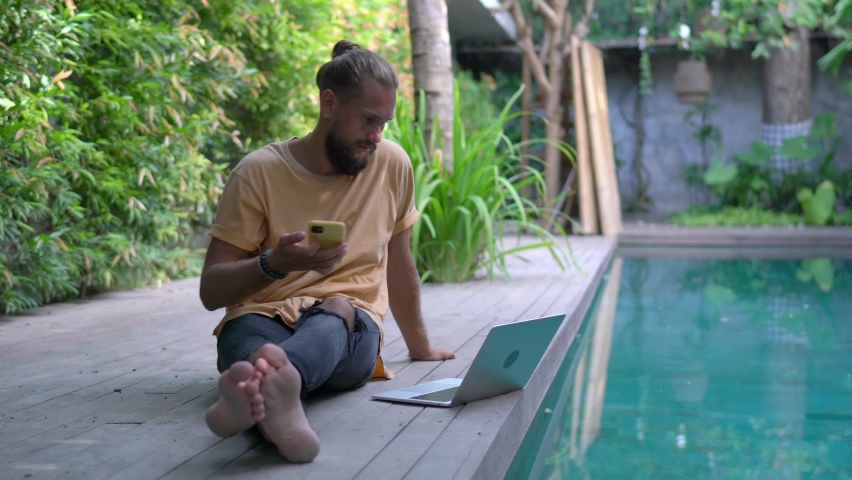 Digital nomad with smartphone connecting to villa internet for doing remote work at pool terrace, skilled male freelancer reading web publication while making online booking via application on netbook - Powered by Shutterstock - Get 15% off with code: PIKWIZARD15