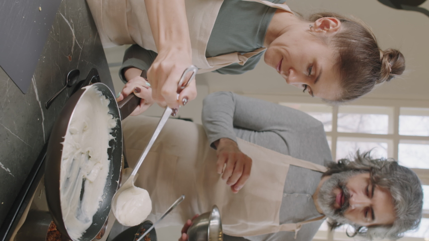 Slow-motion vertical medium shot of man and woman cooking lasagna during cooking class, woman pouring some Bechamel sauce on top of dish