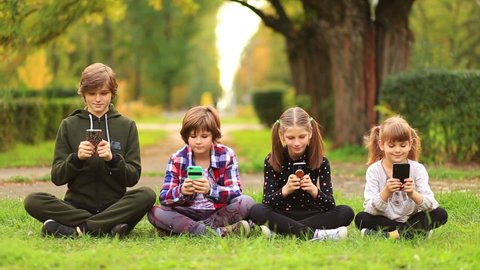 Happy children friend girls group sit with cross legs on grass and playing internet game with mobile smartphone. kids texting message on smartphone outdoor in summer garden. Blogger lifestyle