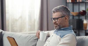 Stylish adult man with glasses sitting at home on couch and watching funny video on tablet while drinking hot drink.