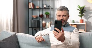 Stylish adult man with gray hair and beard sits at home on couch and talks to friends on video call holding smartphone.