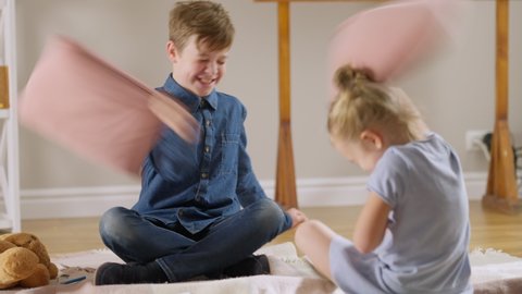 Cheerful boy and girl fighting pillows sitting on carpet in living room and laughing. Positive happy relaxed Caucasian brother and sister or friends having fun indoors at home. Leisure and joy