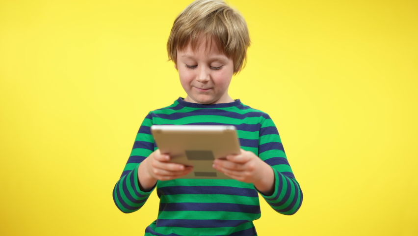Middle shot portrait of cute boy using tablet standing at yellow background. Absorbed Caucasian little gamer gaming online smiling. Happy blond handsome kid enjoying video game | Shutterstock HD Video #1071023653