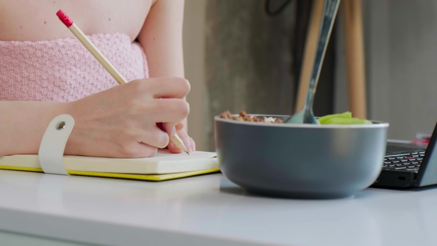 Close up of an young business lady writting notice in her notebook, with a towel on head and eating healthy breaksfast, talking online using a computer with her colleague.